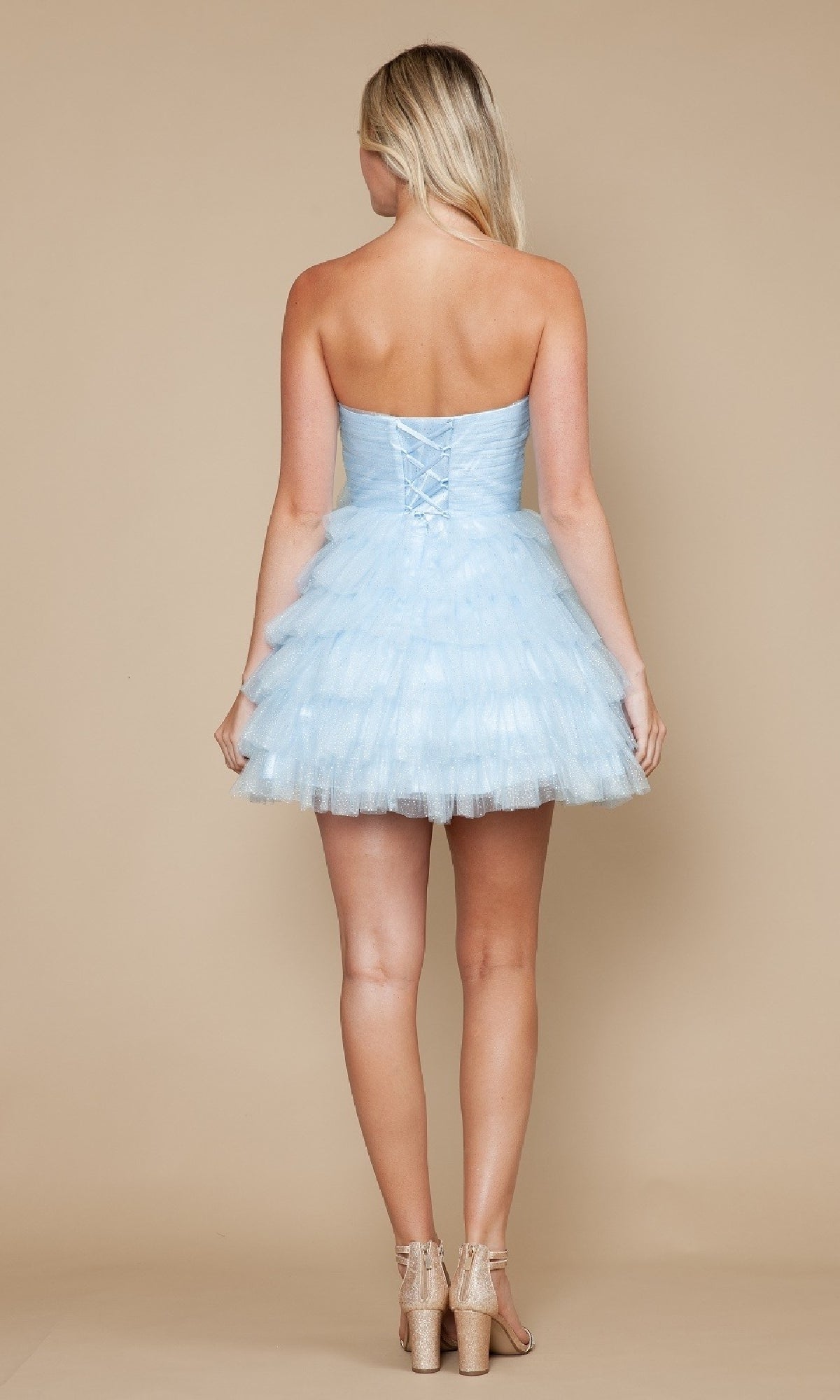 A blonde woman models the Strapless Short Tulle Hoco Dress: Poly USA 9912—a light blue, layered mini dress with ruffles and lace-up back—while posing against a beige background.