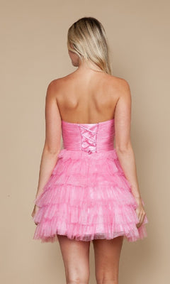 A person with long blonde hair is shown from the back wearing the Strapless Short Tulle Hoco Dress: Poly USA 9912, a pink strapless party dress with lace-up corset detail and playful ruffles.