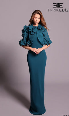 A woman wearing the Tarik Ediz 99134 long formal dress in Atlantic Blue, featuring a high neck and 3D floral appliqué on the shoulders, stands against a plain background. The Tarik Ediz logo appears in the top right corner.