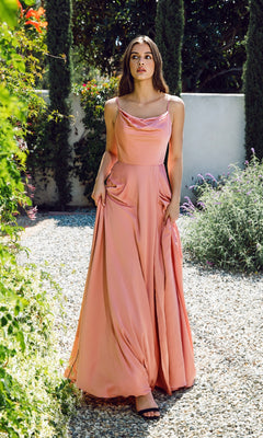 A woman stands outdoors on a gravel path, surrounded by lush greenery, wearing the Eureka 9916 Cowl-Neck Long Satin Formal Dress in peach with thin straps and black sandals.
