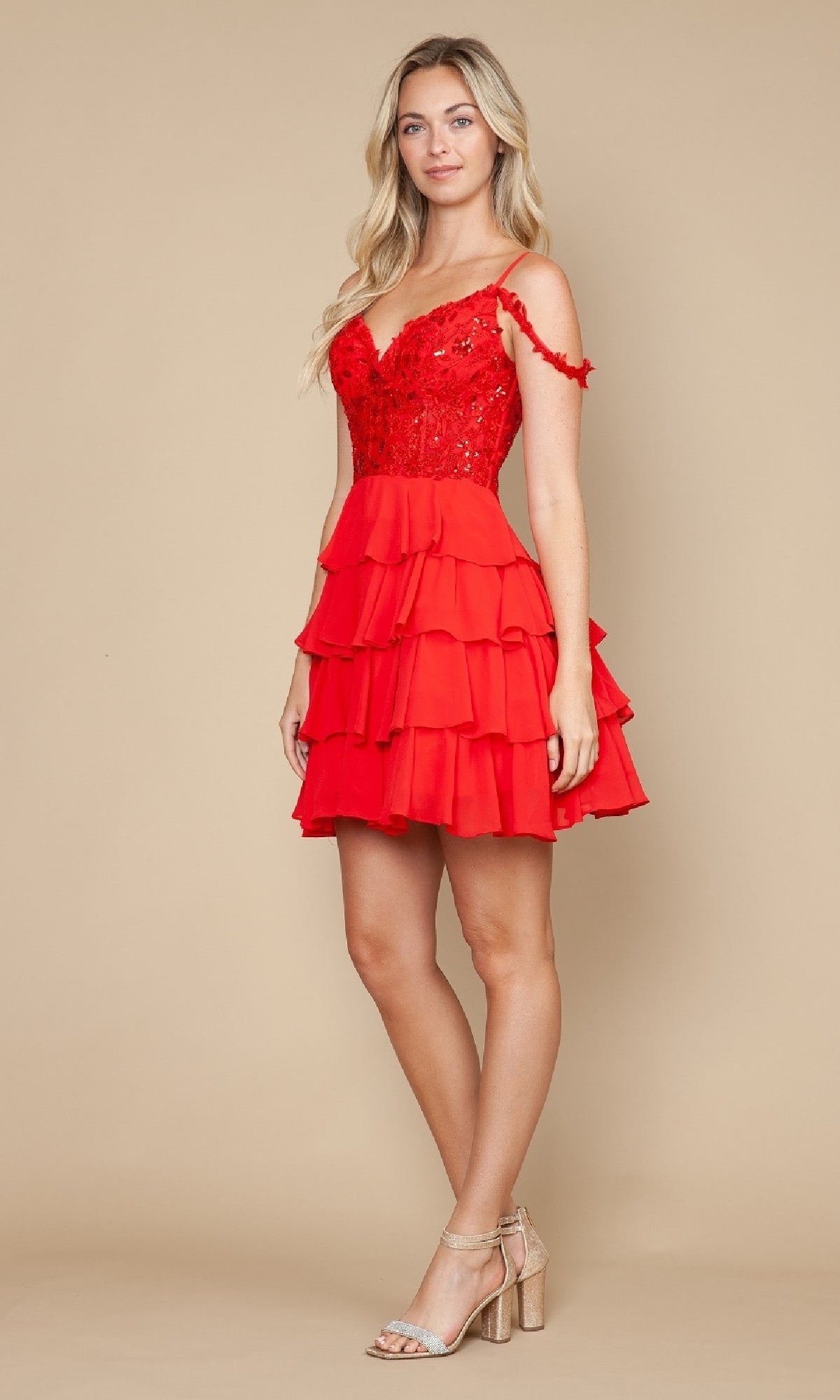 A woman poses in the Poly USA 9920 short homecoming dress, featuring red ruffles, an off-the-shoulder lace bodice, and a chiffon A-line skirt, styled with nude block-heeled sandals against a beige background.