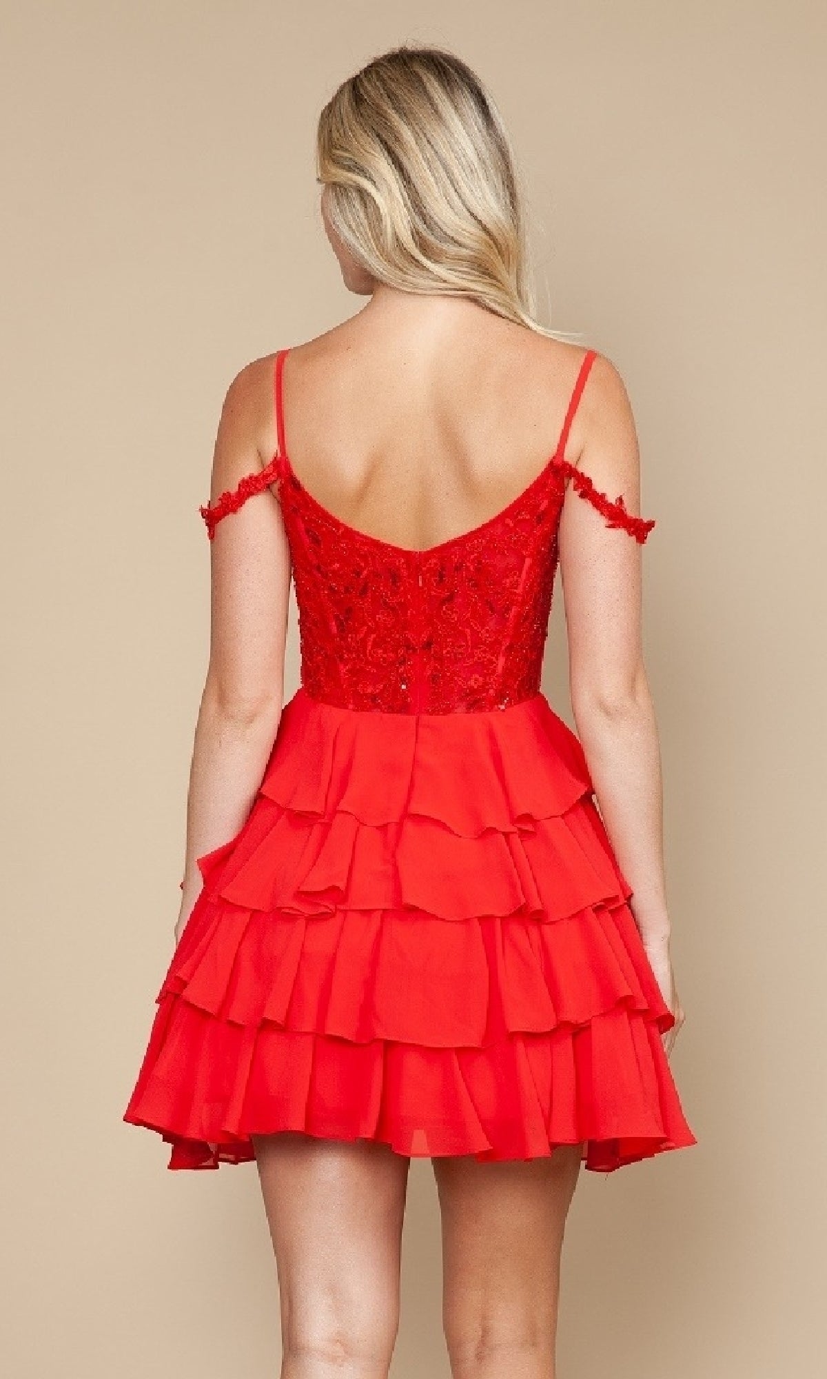 A woman faces away wearing the Poly USA 9920 short homecoming dress, featuring a red fit-and-flare silhouette, fitted lace bodice, off-the-shoulder straps, and tiered ruffled skirt against a plain beige background.