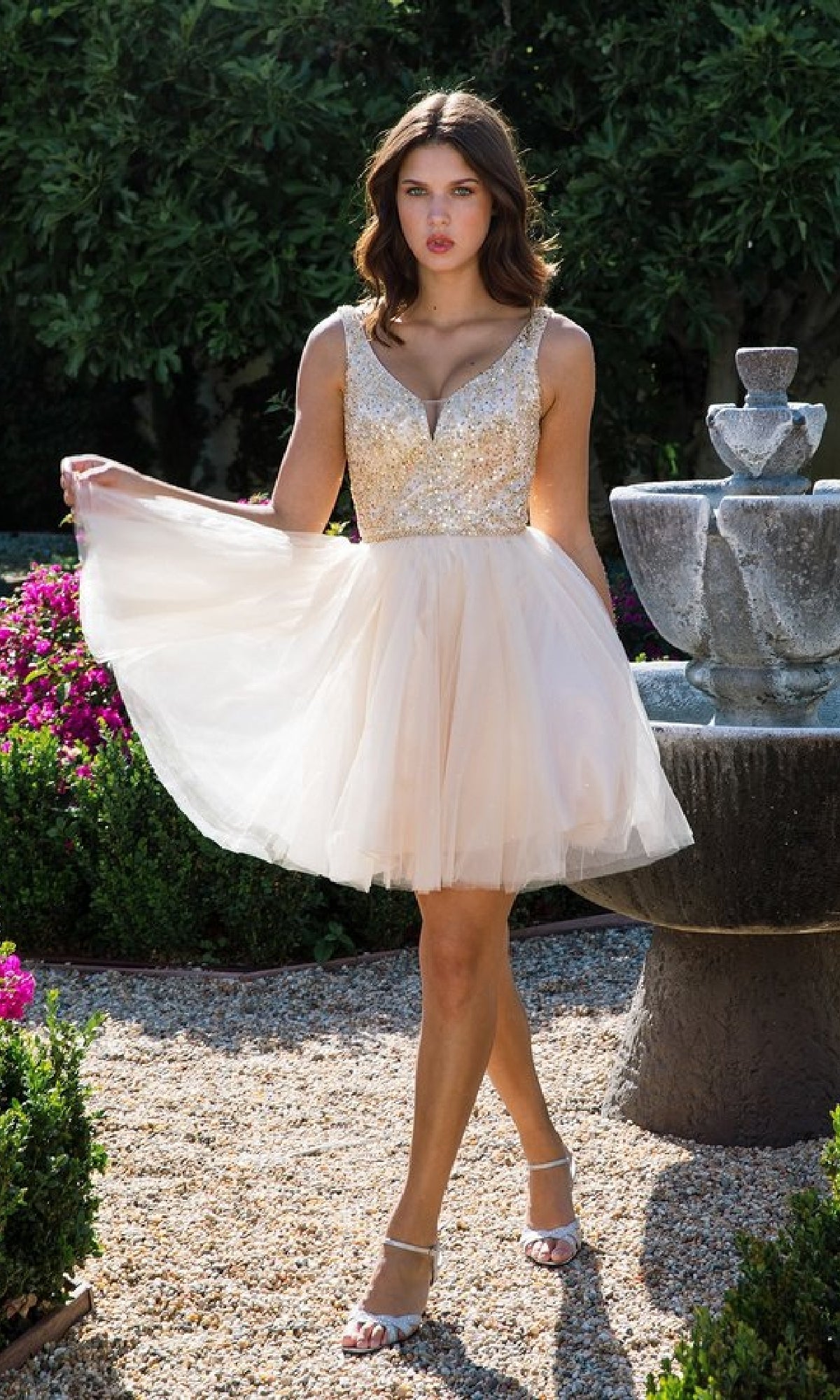 A woman in the Glitter-Bodice Short A-Line Hoco Dress (Eureka 9926), featuring a sleeveless, sequin bodice, stands outdoors near a stone fountain surrounded by lush greenery.