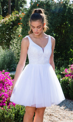 A young woman wears the Glitter-Bodice Short A-Line Hoco Dress: Eureka 9926, featuring a sleeveless white bodice and tulle skirt, as she stands in a blooming garden surrounded by lush greenery.