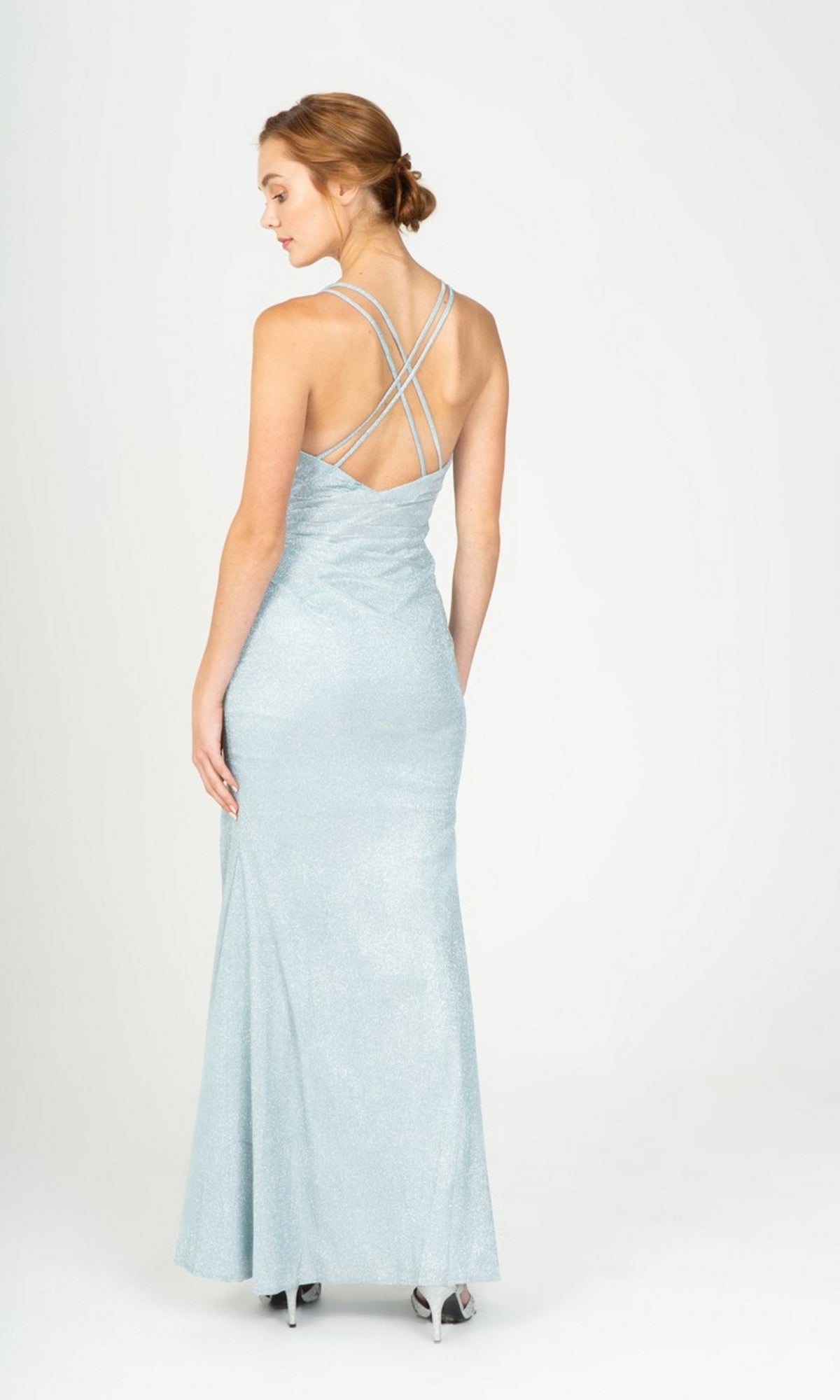 A woman stands facing away in the Long Prom Dress: Eureka 9933, a shimmering light blue glitter jersey gown with crisscross straps and silver heels against a plain background.