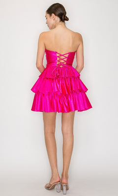 A woman stands facing away, wearing the Poly USA 9944 short homecoming dress—a strapless hot pink fit-and-flare style with a lace-up back—paired with clear high-heeled sandals.