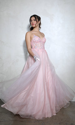 A woman stands against a textured, light-colored wall wearing the Lace-Up Corset Long Prom Ball Gown: Eureka 9960, a light pink A-line dress with thin straps and floral embellishments.