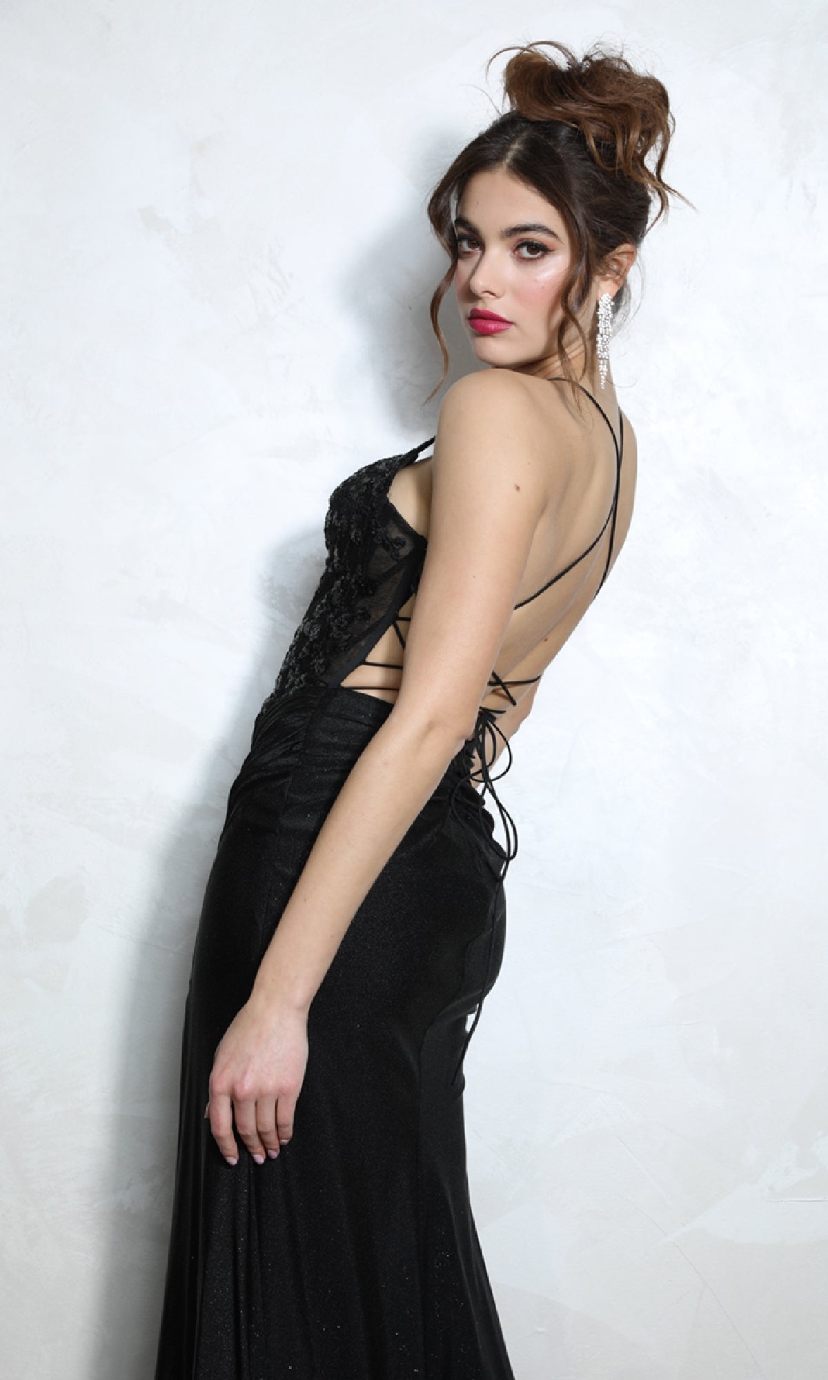A woman in the Floral-Bodice Long Glitter Prom Dress: Eureka 9969 stands against a white textured wall, looking over her shoulder with a neutral expression.