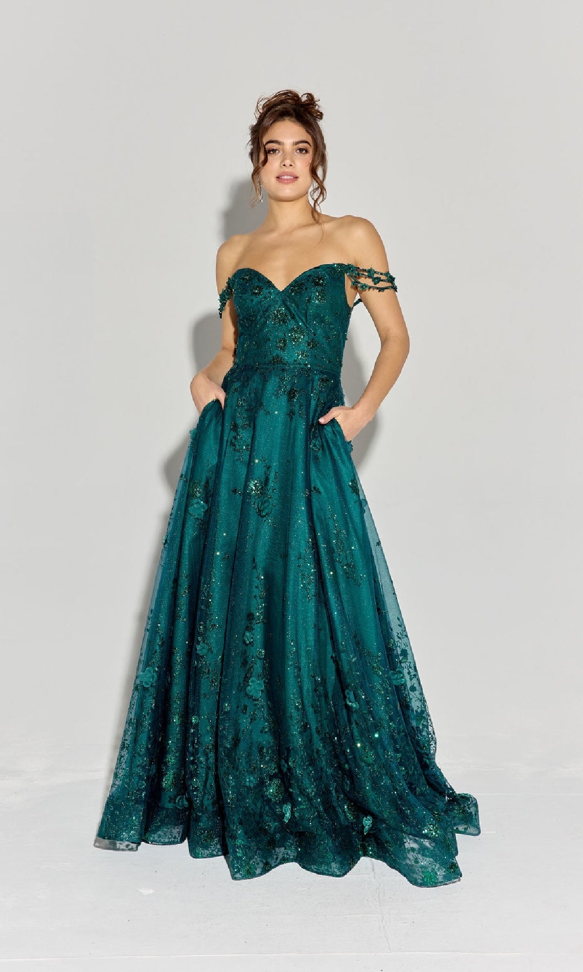 A woman stands against a plain background wearing the Long Glitter-Tuller A-Line Prom Dress: Eureka 9980, an off-the-shoulder, floor-length gown featuring floral embellishments, side pockets, and elegant glitter-tulle details.
