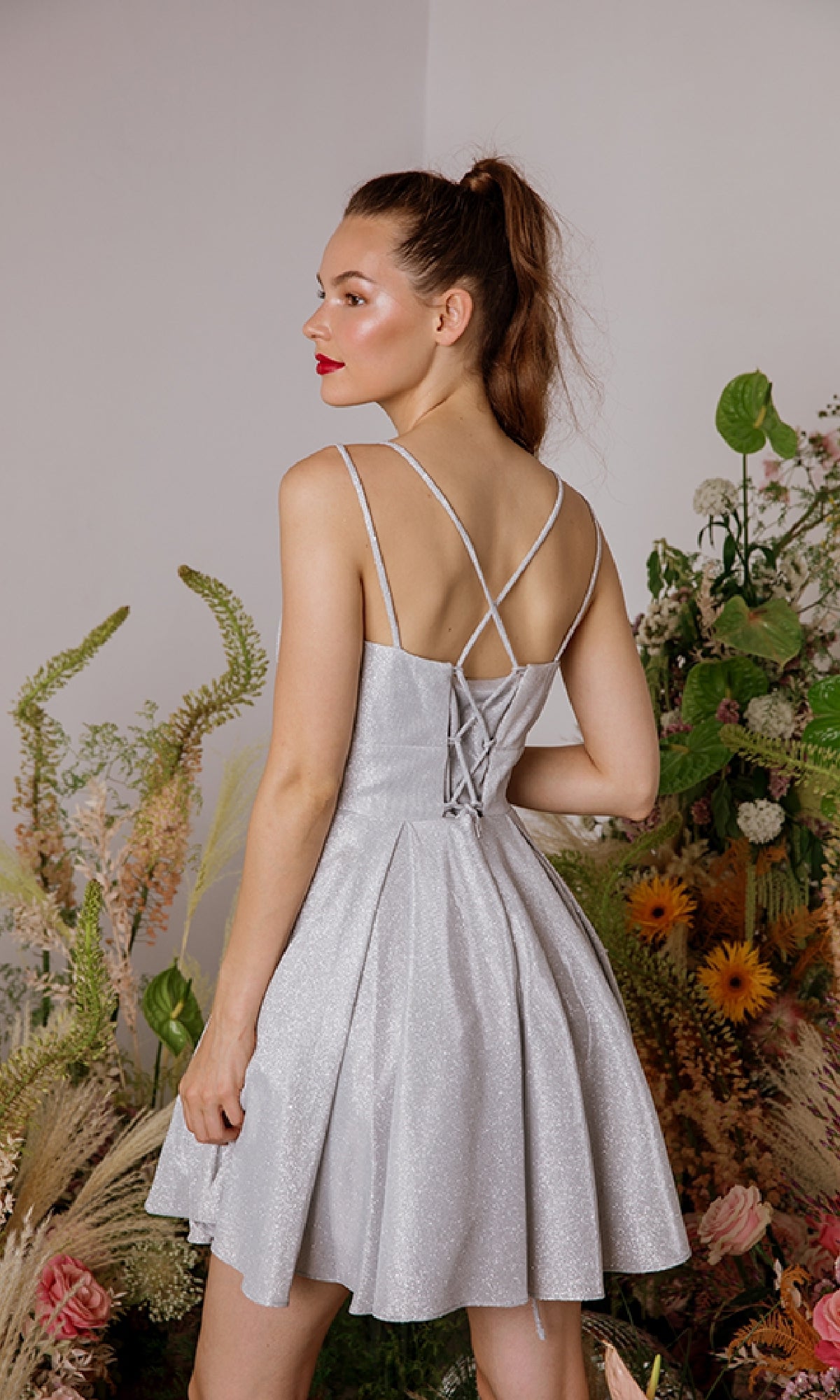 A woman in the Eureka 9988 Lace-Up Short Glitter Hoco Dress, featuring a sleeveless crisscross-back design in light silver, stands among green plants and flowers with her back to the camera.