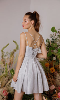 A woman in the Eureka 9988 Lace-Up Short Glitter Hoco Dress, featuring a sleeveless crisscross-back design in light silver, stands among green plants and flowers with her back to the camera.