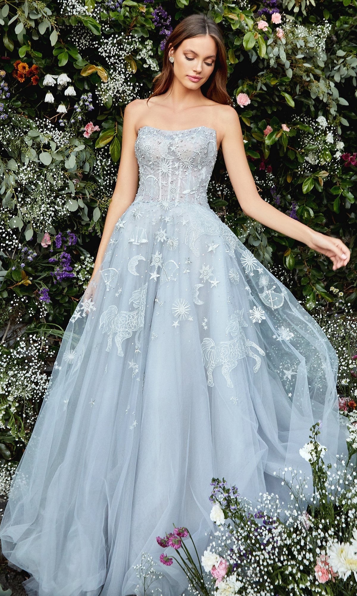 A woman stands outdoors in front of greenery, wearing the Constellation-Print Strapless Prom Ball Gown A0890, a light blue gown with celestial and animal embroidery and a tulle skirt.