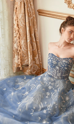 A woman wearing the Constellation-Print Strapless Prom Ball Gown A0890 sits by an ornate curtain and gold-trimmed wall.
