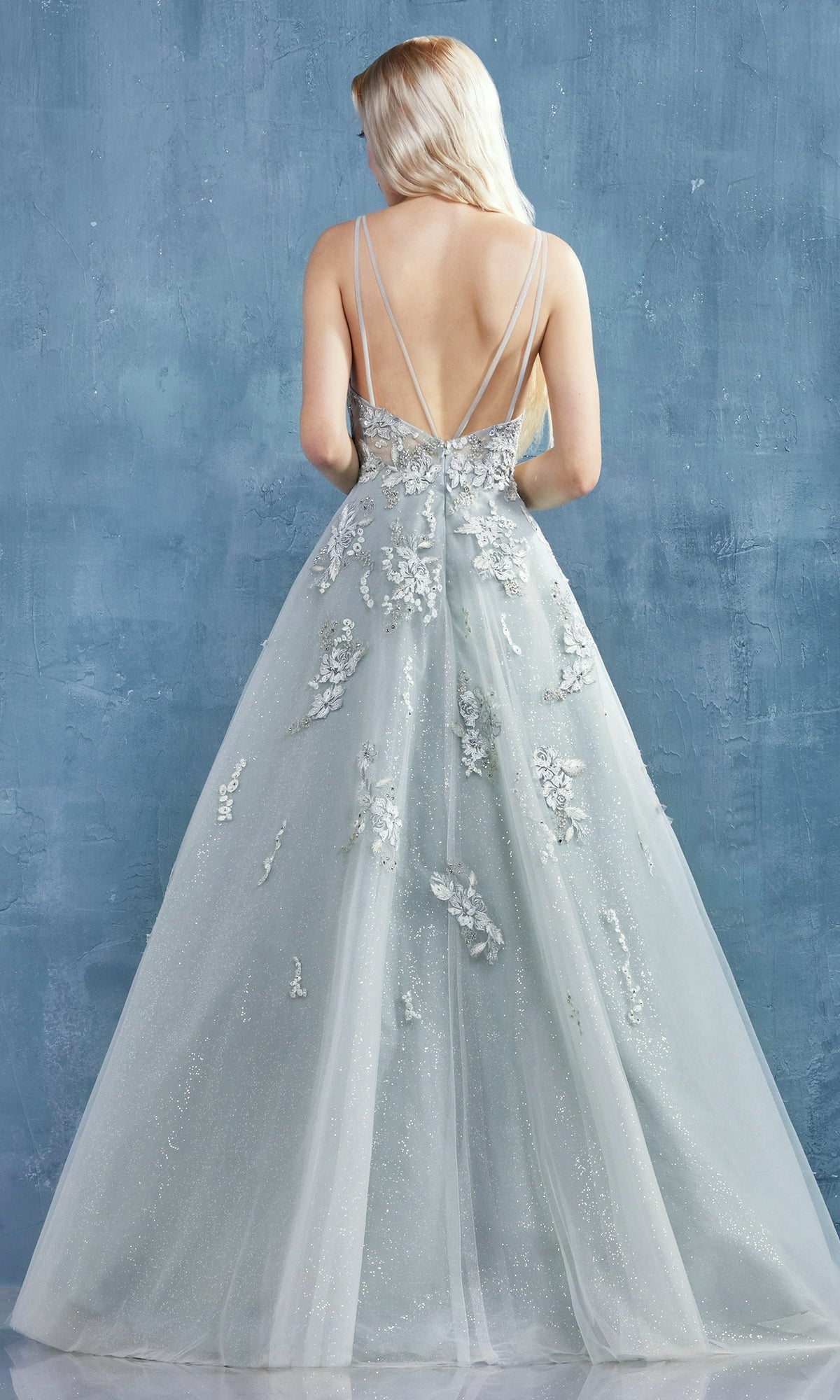 A person with long, light blonde hair wears the Sage Green Long Glitter Prom Dress A0892 with floral appliqués and thin straps, facing away against a blue textured background.