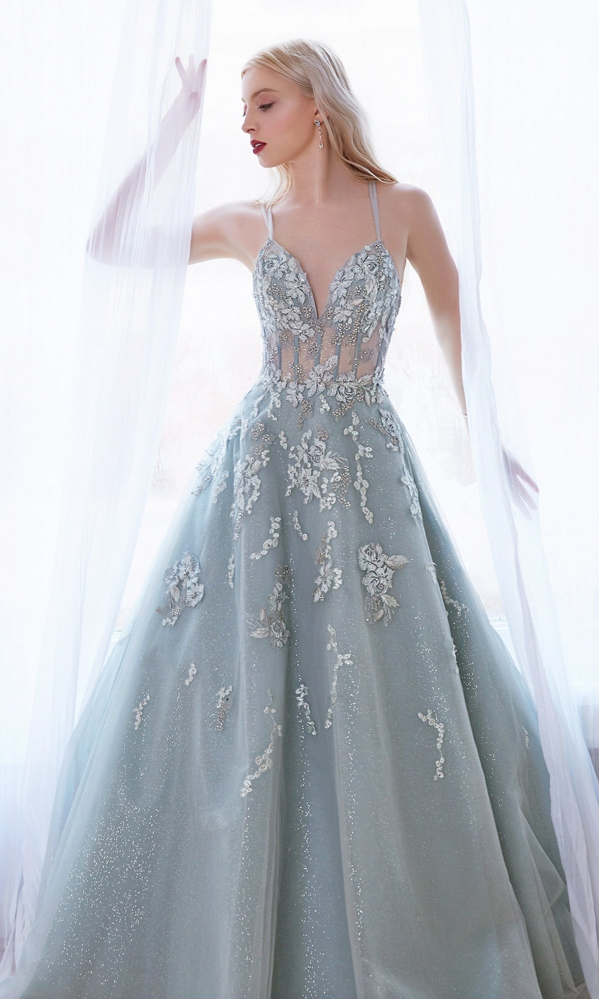 A woman in a sage green long glitter prom dress (A0892) stands by a sheer white curtain, one arm raised, gazing to the side as her gown shimmers.