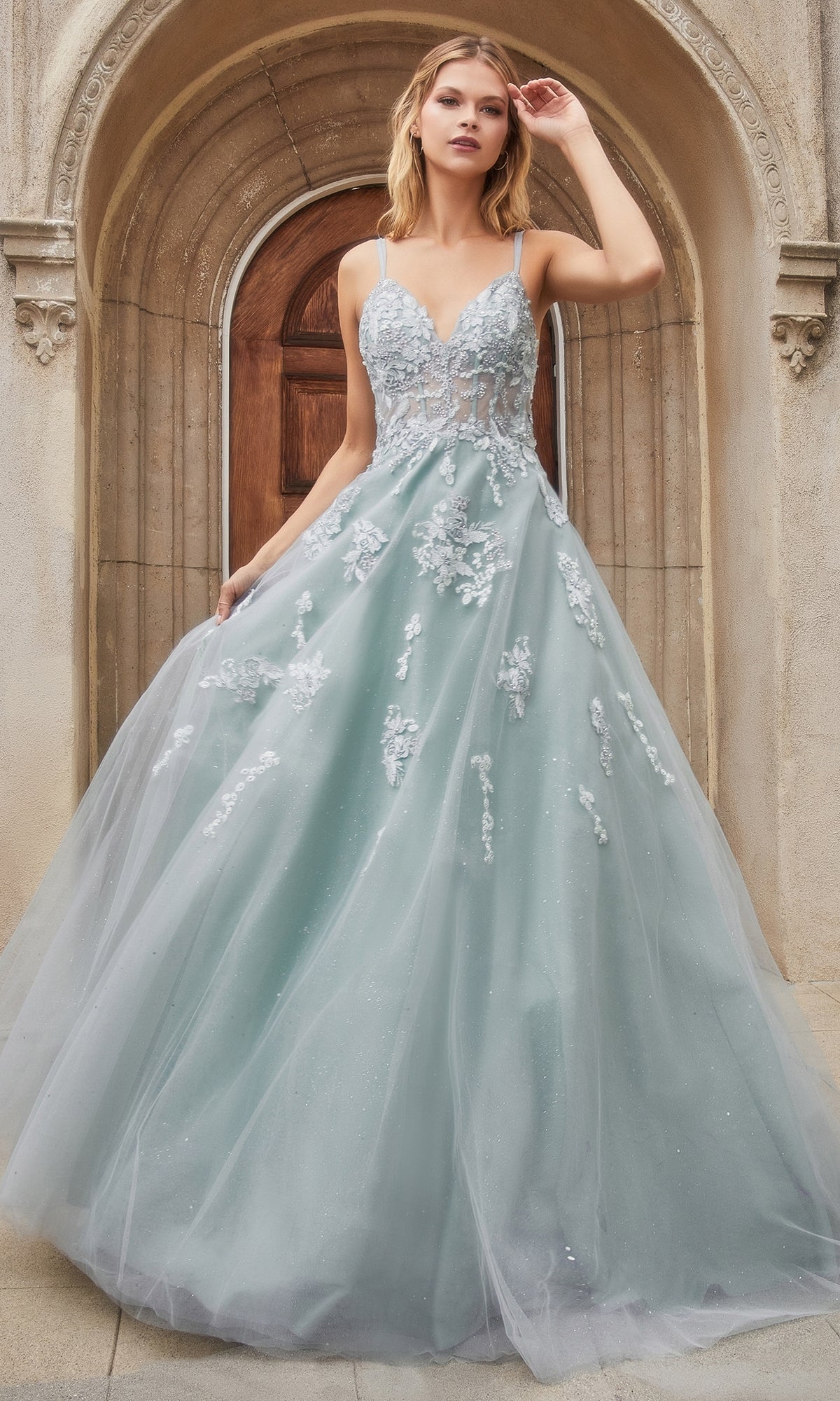 A woman in a light blue gown with floral appliqués stands before an arched doorway, capturing the elegance of the Sage Green Long Glitter Prom Dress A0892, with one hand raised near her head.