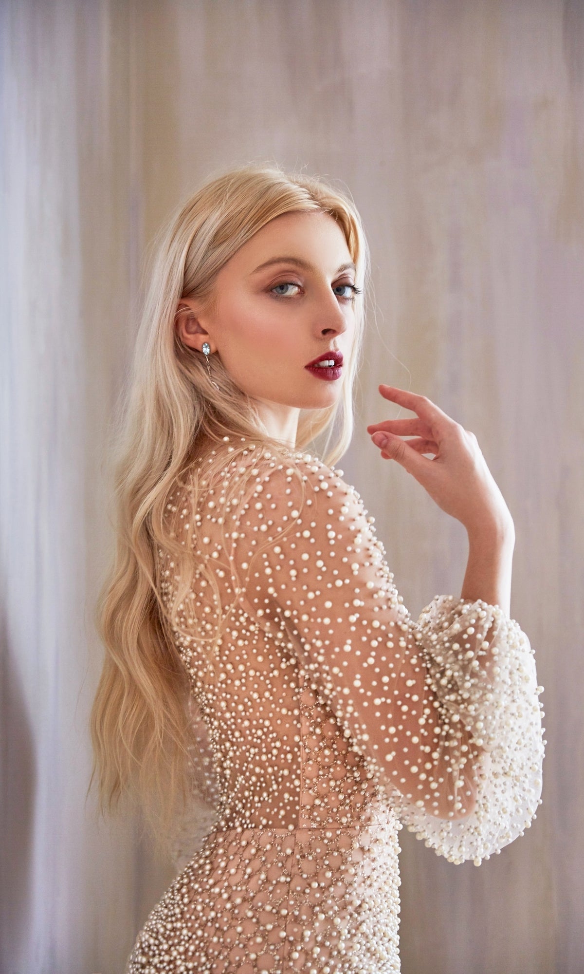 A woman with long blonde hair models the Pearl-Embellished Long Sleeve Formal Dress CLA0997, looking over her shoulder against a soft, neutral background.