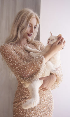 A woman with long blonde hair, dressed in the Pearl-Embellished Long Sleeve Formal Dress CLA0997, holds a white cat near a light-colored wall.