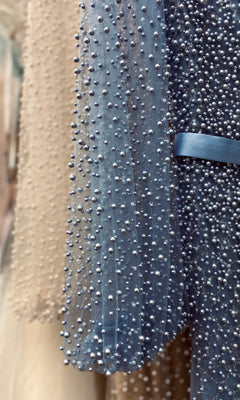 Close-up of the Pearl-Embellished Long Sleeve Formal Dress CLA0997 in sheer blue with a satin waistband and delicate pearl details; a beige version with similar beading is seen in the background.