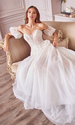 A woman in the Puff-Sleeve Long White Wedding Dress A1014 with off-the-shoulder lace, removable puff sleeves, and a full tulle skirt sits on a beige sofa in an elegant room with wood floors.