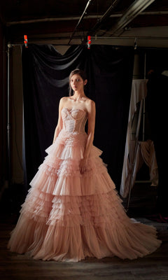 A woman stands before a black backdrop in a dimly lit room, wearing the Strapless Tiered Long Prom Ball Gown A1017, featuring layers of pink ruffles.