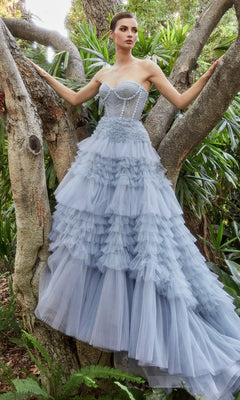 A woman wearing the Strapless Tiered Long Prom Ball Gown A1017 in light blue stands outdoors among tree branches and lush greenery, with delicate ruffles accentuating her look.