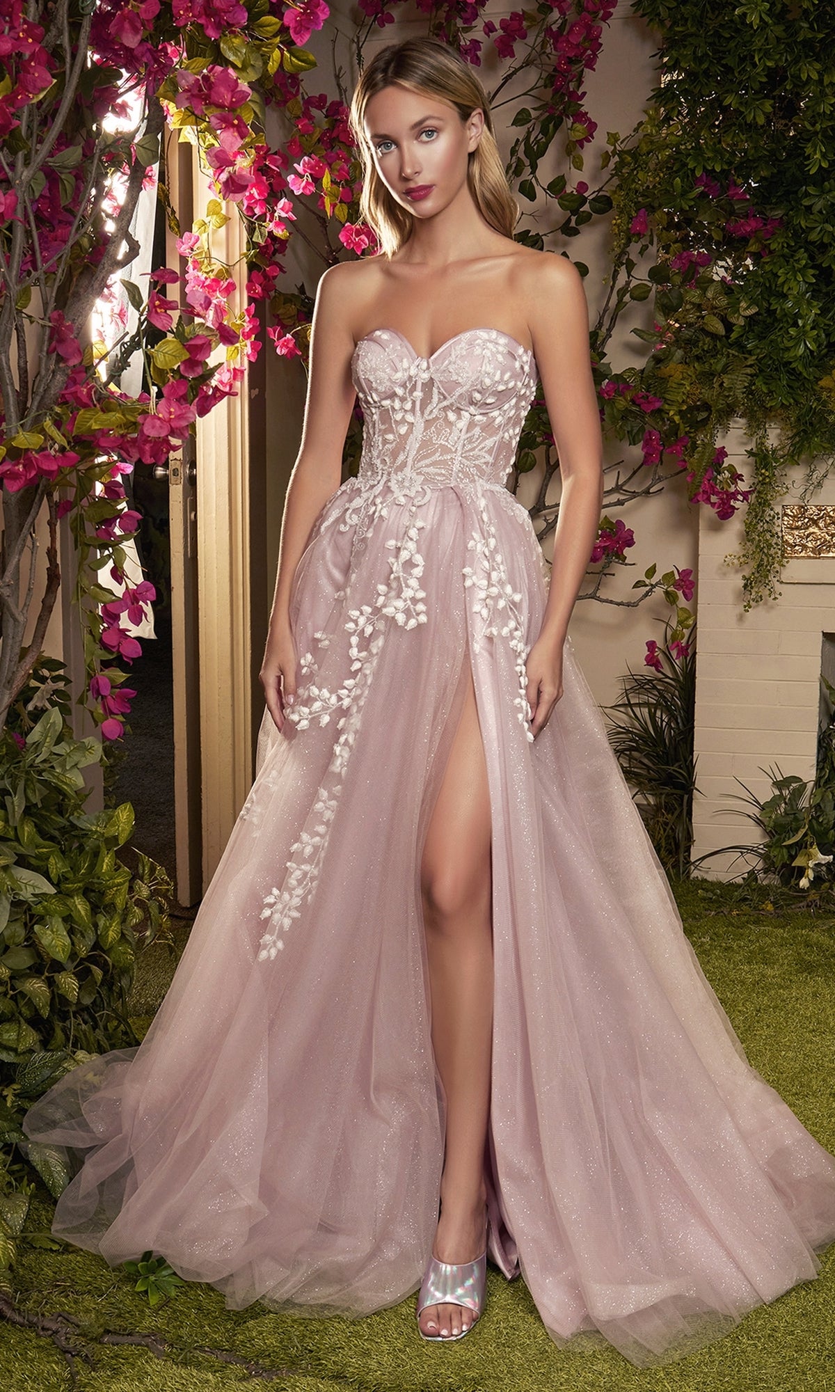 A woman stands outdoors in front of flowers, wearing the Strapless Sweetheart Prom Ball Gown A1029 featuring floral appliqué, a fitted bodice, high slit, and paired with metallic open-toe heels.