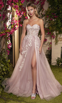 A woman stands outdoors in front of flowers, wearing the Strapless Sweetheart Prom Ball Gown A1029 featuring floral appliqué, a fitted bodice, high slit, and paired with metallic open-toe heels.