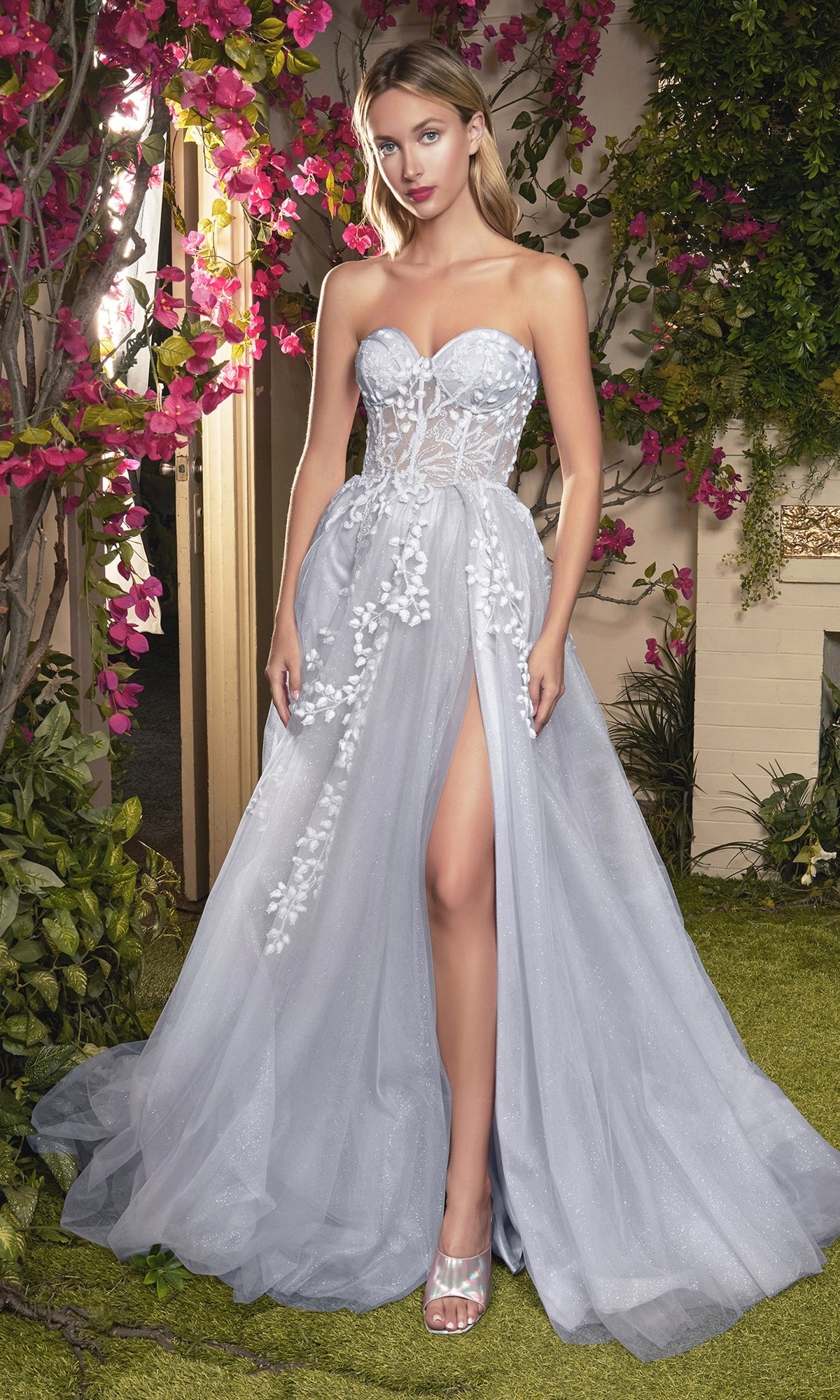 A woman in the Strapless Sweetheart Prom Ball Gown A1029, featuring floral appliqués and a high slit, stands in a garden with pink flowers and greenery.