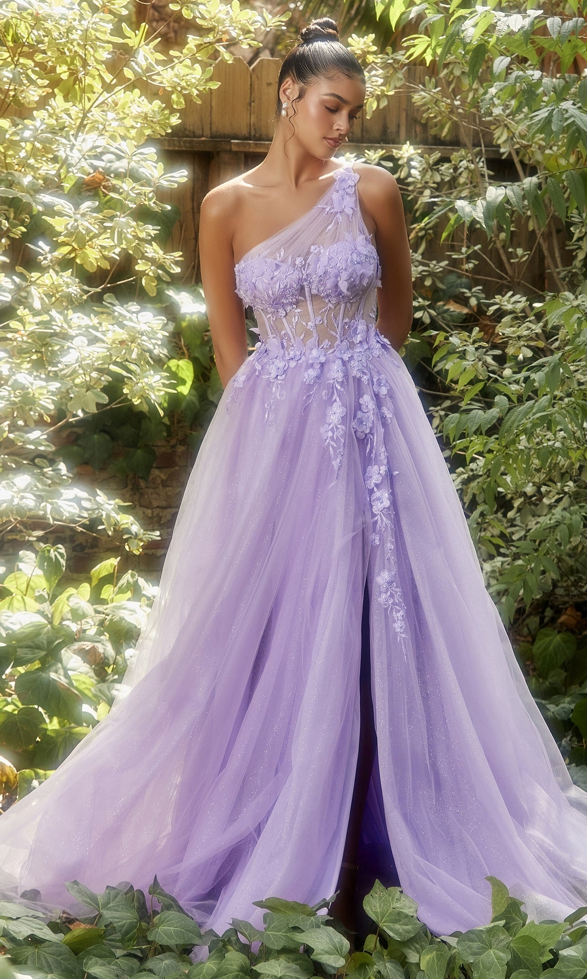 A woman stands in a sunlit garden, surrounded by greenery, wearing the Sheer-Bodice One-Shoulder Prom Ball Gown A1140 in lilac with floral applique and a sheer corset bodice.