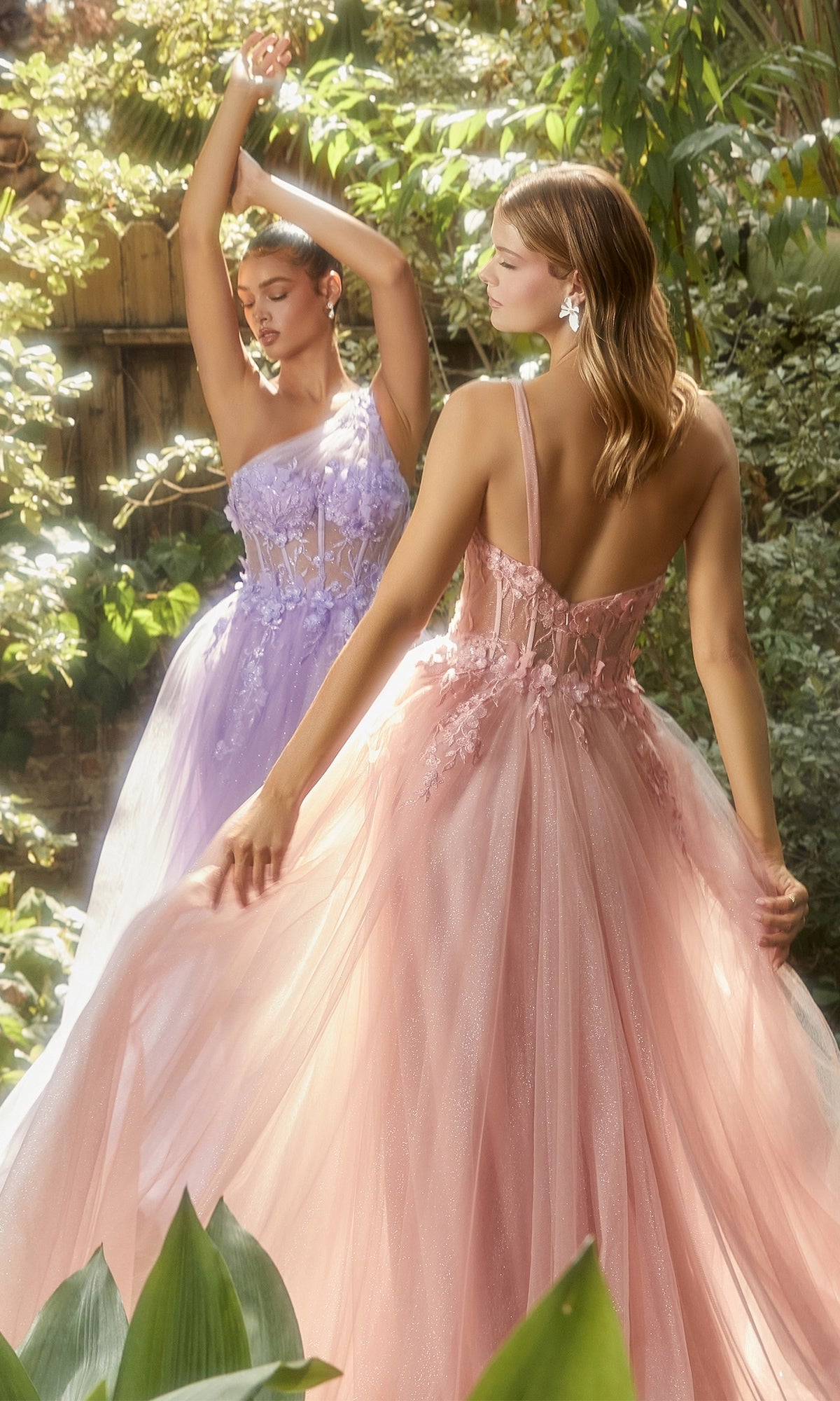Two women stand in a sunlit garden wearing Sheer-Bodice One-Shoulder Prom Ball Gown A1140, floor-length with pastel floral applique details—one in lavender, one in blush pink—as sunlight filters through lush greenery.