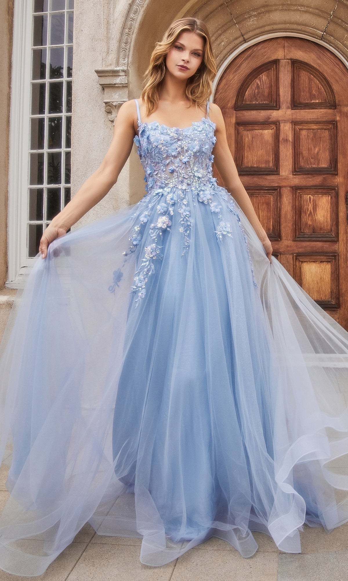 A woman stands outside by a wooden door, wearing the Flower-Embellished Long Tulle Prom Ball Gown A1142—a floor-length light blue gown with a sheer waist and 3D floral accents on its elegant, flowing tulle skirt.