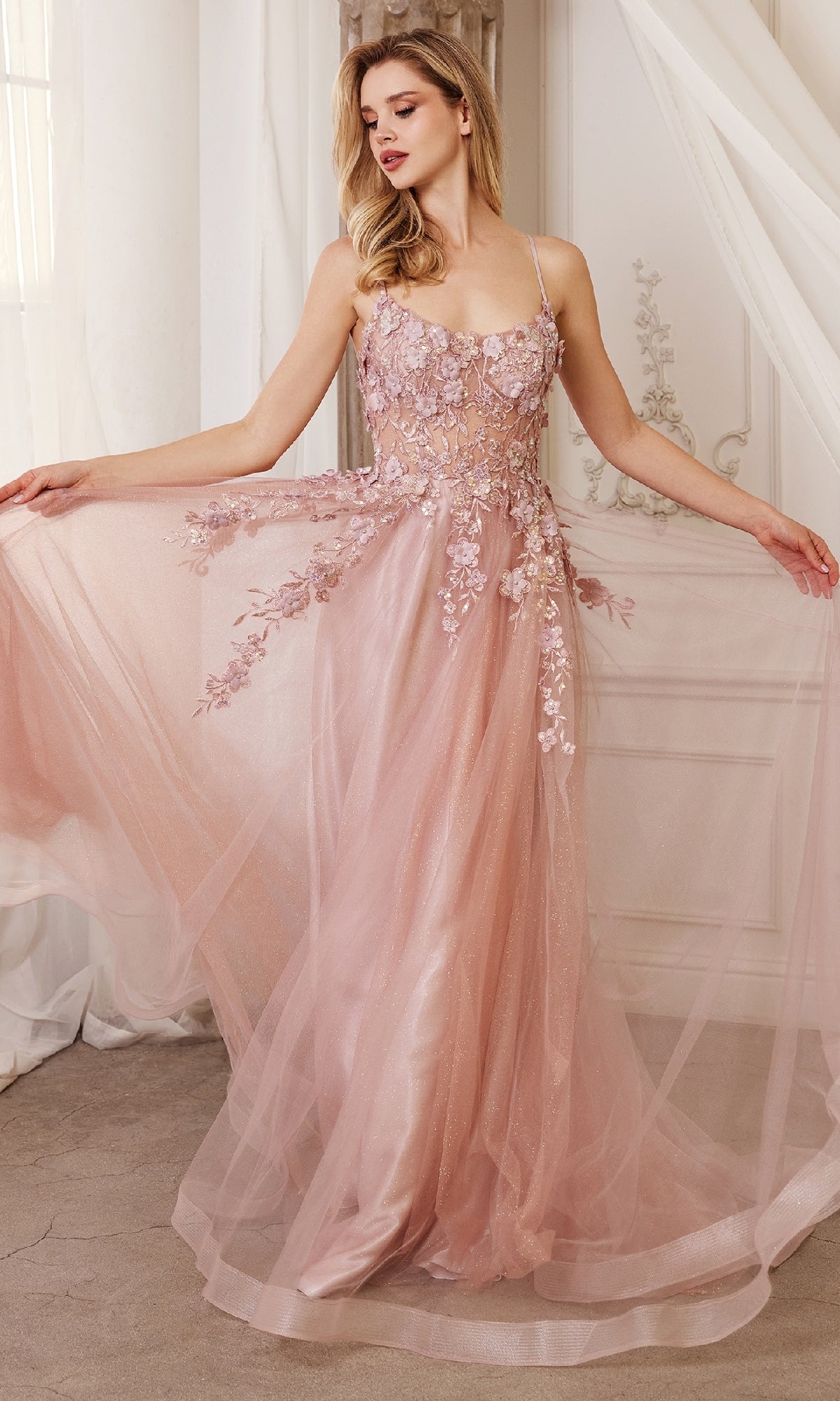 A woman stands indoors holding out the sides of the Flower-Embellished Long Tulle Prom Ball Gown A1142, a flowing blush pink dress with 3D floral details on the bodice and skirt and a sheer waist.