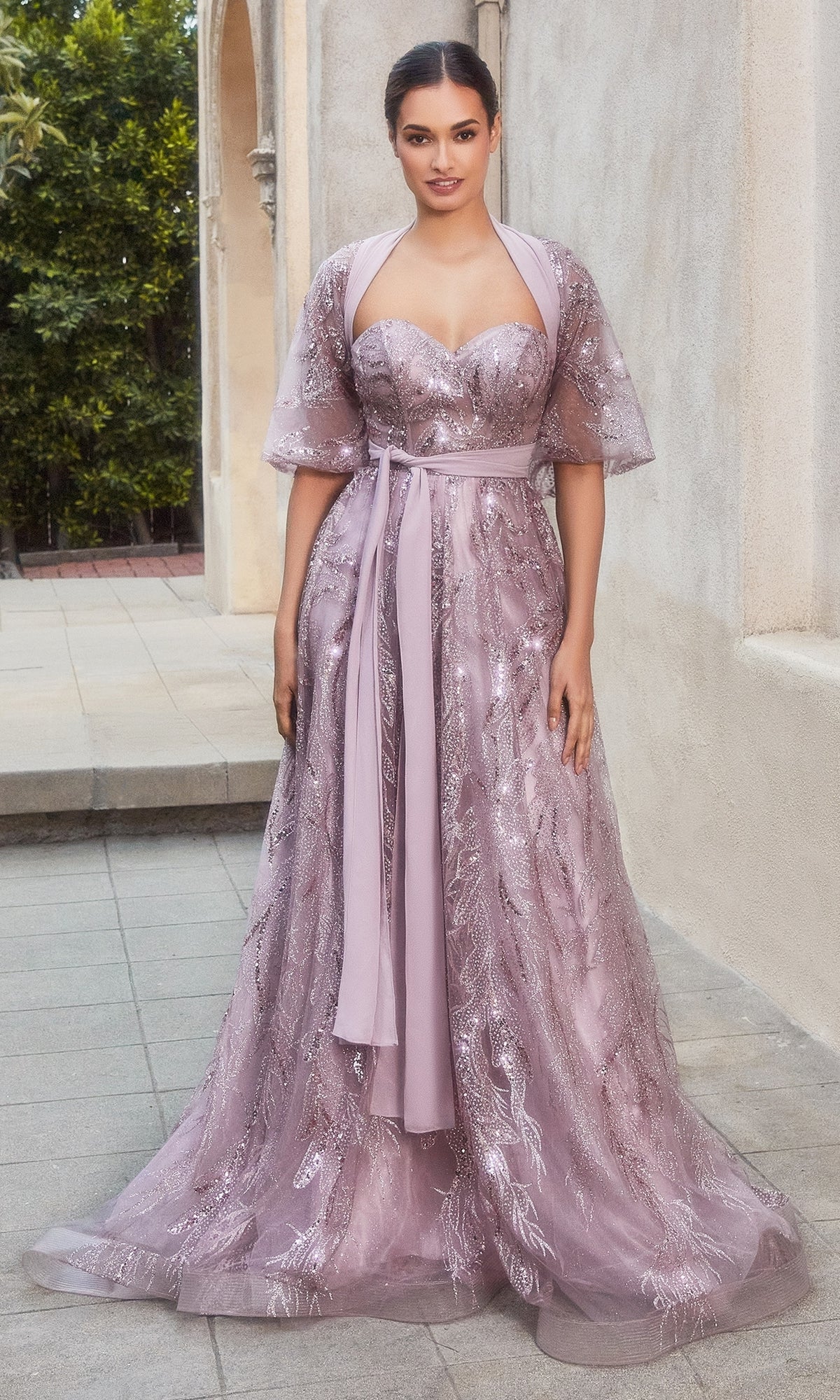 Outdoors, a woman models the Strapless Long Prom Ball Gown with Capelet A1174—a floor-length mauve pink beaded formal dress featuring a sheer, sparkling overlay and a matching sash belt.