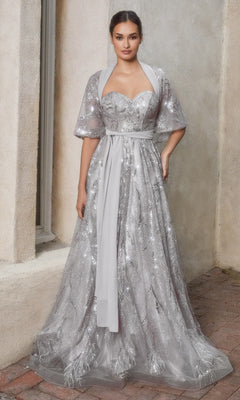 A woman stands against a textured wall wearing the Strapless Long Prom Ball Gown with Capelet A1174—a floor-length, silver sequined gown featuring a capelet and elegant detailing.