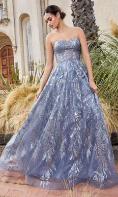 A woman wearing the Strapless Long Prom Ball Gown with Capelet A1174 stands outdoors near tall grasses and a palm tree. The elegant silver-blue gown adds sophistication to the tranquil setting.
