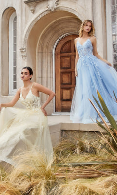 Two women in long, sleeveless tulle dresses—one in the Floral-Embroidered Long A-Line Prom Gown A1191 in pale yellow, the other in a light blue v-neck evening gown—pose before an arched stone doorway framed by ornamental grass.