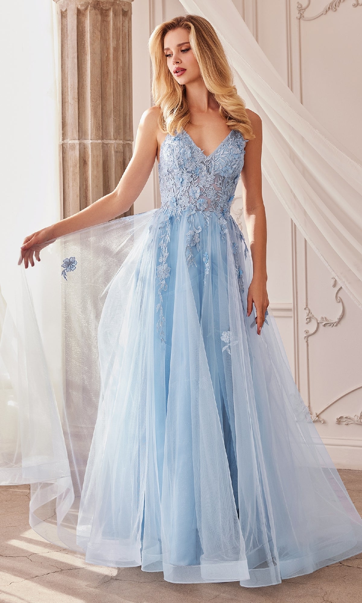 Indoors, a woman wears the Floral-Embroidered Long A-Line Prom Gown A1191, featuring light blue lace and tulle with floral embroidery, holding a section of the floor-length skirt to the side.