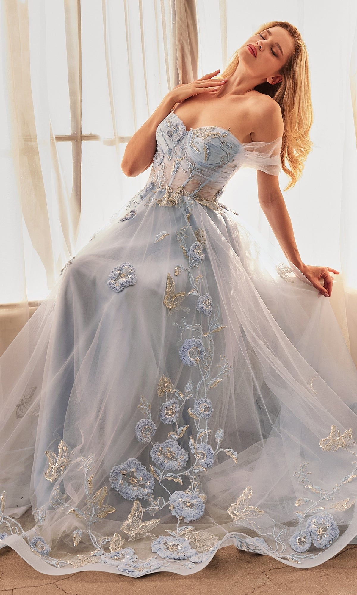 Wearing the Andrea and Leo A1246 Long Blue Prom Ball Gown, a woman sits on the floor before sheer curtains with her eyes closed and head tilted back.