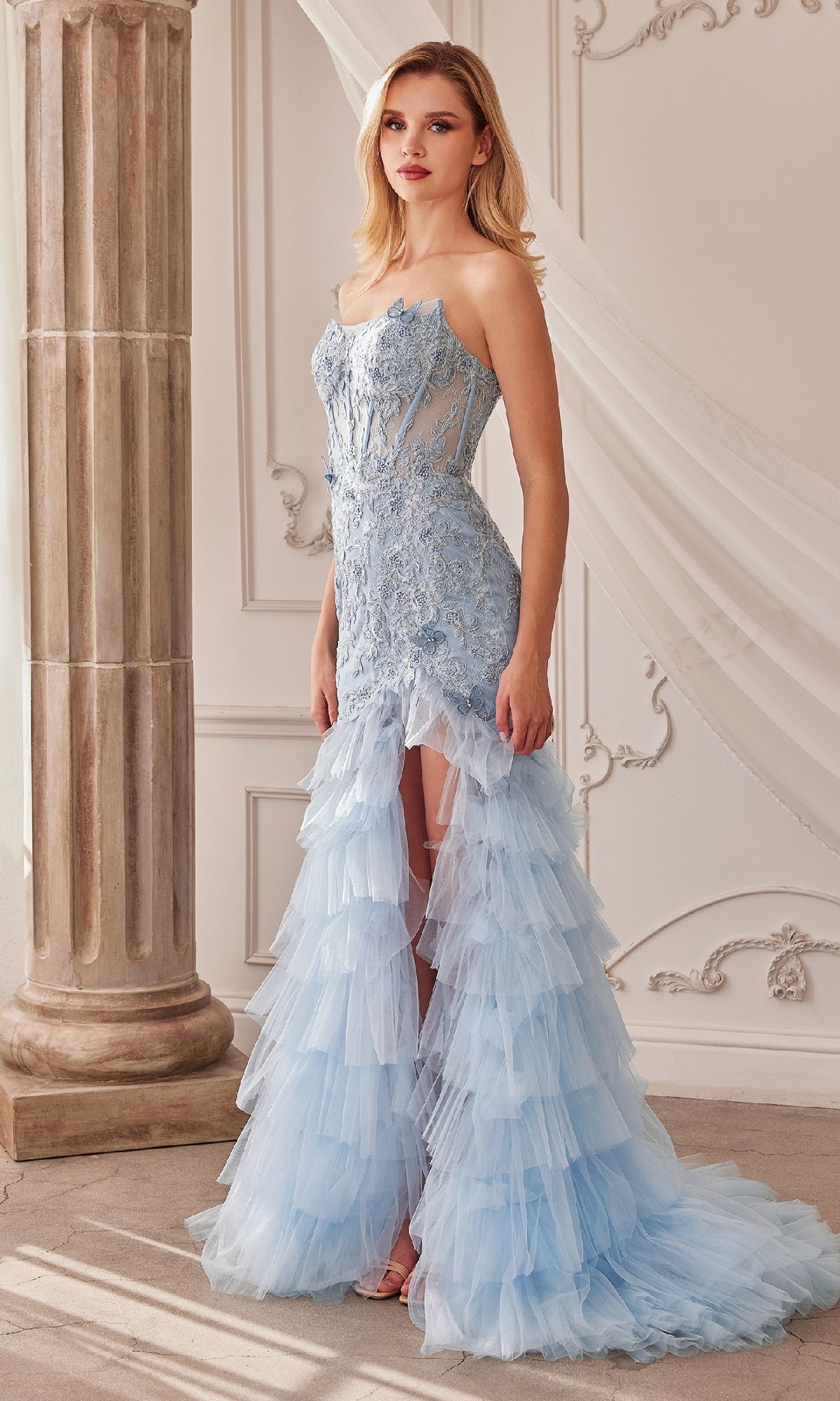 A woman in the Strapless Fitted Ruffle Prom Dress A1255, featuring a lace bodice and ruffle tulle skirt, stands indoors beside a column and draped fabric.