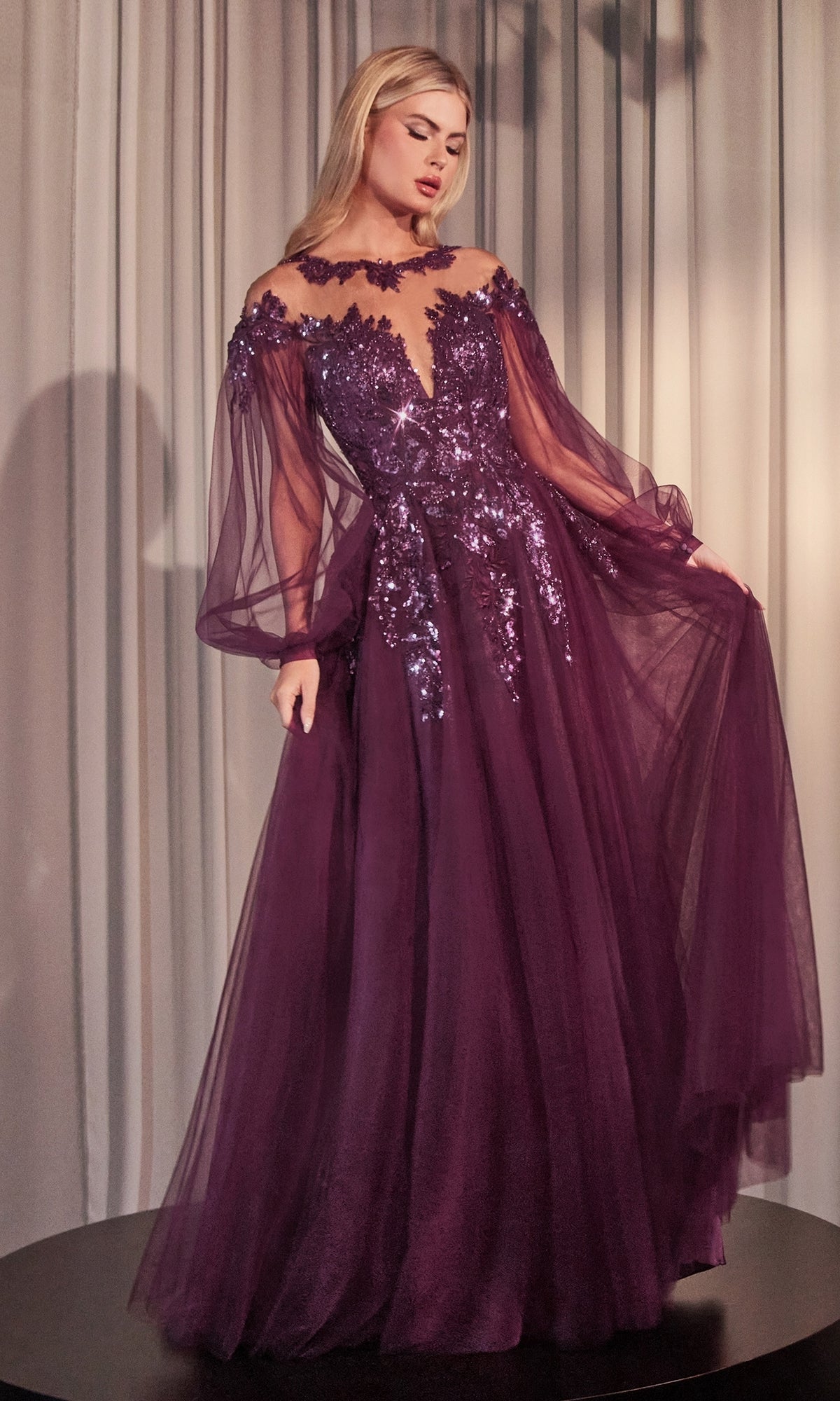 A woman wears the Sheer Long Sleeve Long Formal Dress A1263, a floor-length purple gown with off-the-shoulder design, sheer sleeves, and sequin embellishments, set against light-colored draped curtains.