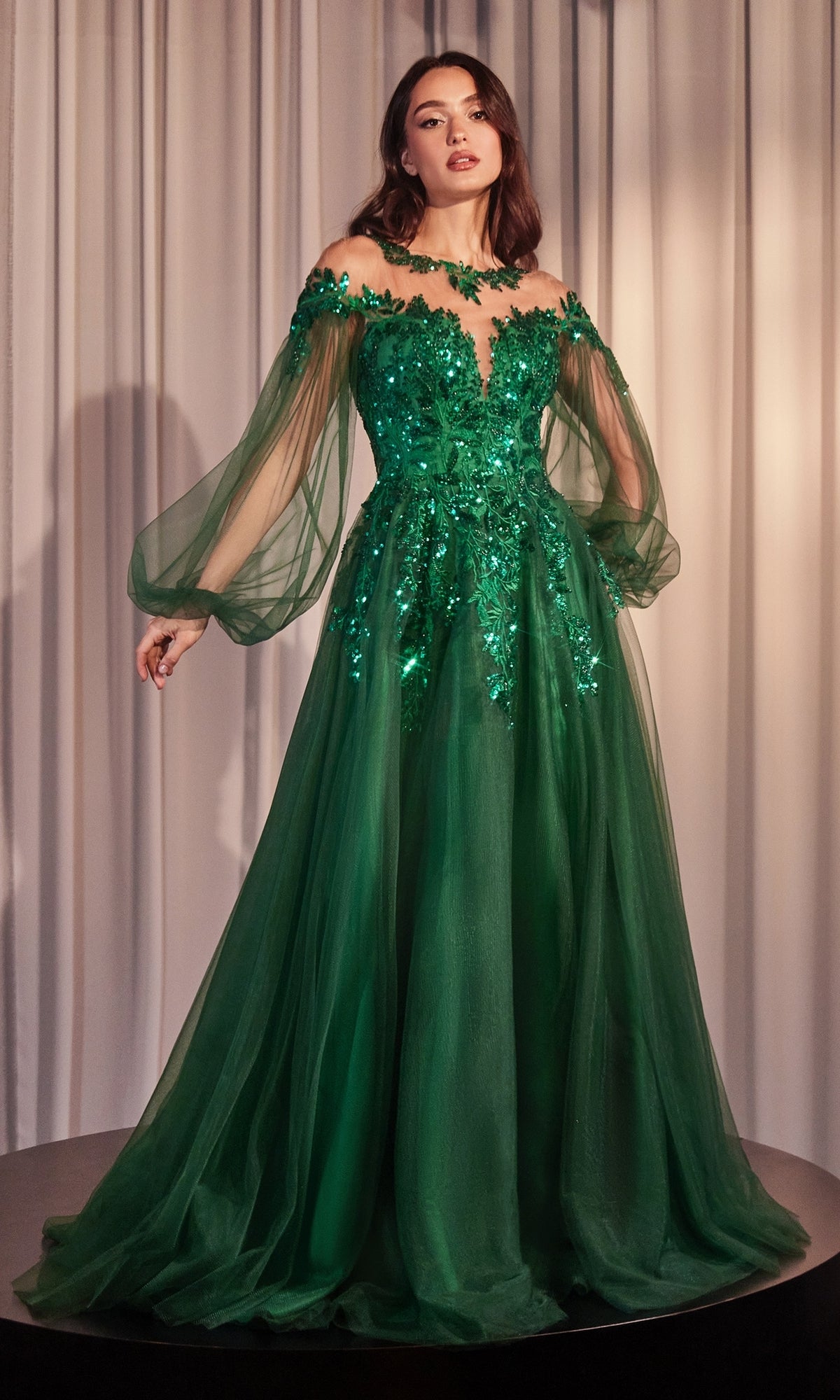A woman stands before white curtains wearing the Sheer Long Sleeve Long Formal Dress A1263—an off-the-shoulder, floor-length green gown with floral embellishments and sheer, voluminous long sleeves.