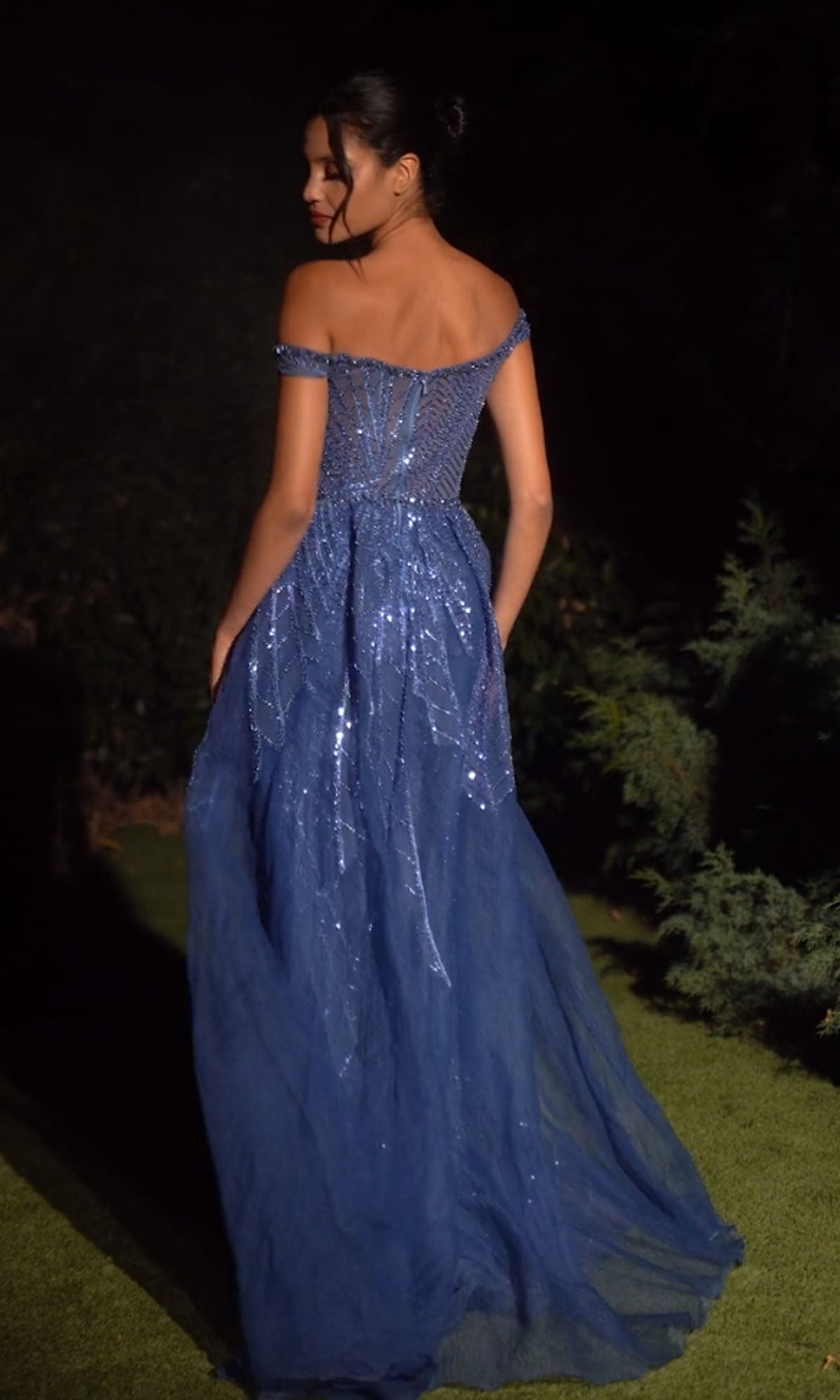 A woman stands outdoors at night wearing the Long Prom Dress A1278 by Andrea and Leo, a blue off-the-shoulder gown with sparkling sequins and a sheer, floor-length overskirt.
