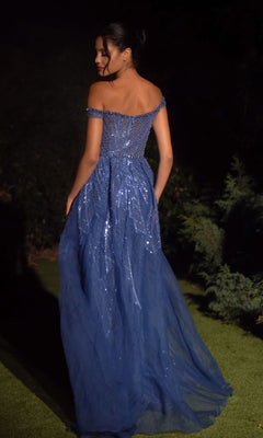 A woman stands outdoors at night wearing the Long Prom Dress A1278 by Andrea and Leo, a blue off-the-shoulder gown with sparkling sequins and a sheer, floor-length overskirt.