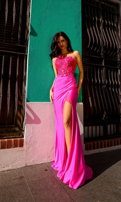 A woman in the One-Shoulder Bright Prom Gown: Nox Anabel A1317 with a beaded sheer bodice and thigh-high slit stands against a turquoise and pink wall in sunlight.