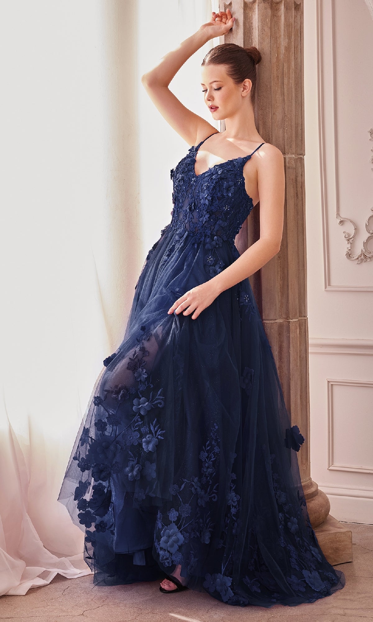 A woman in the Andrea and Leo A1326 Long Glitter Prom Ball Gown stands by a pillar, holding part of her navy blue sleeveless A-line dress with floral appliqué details and looking to the side.