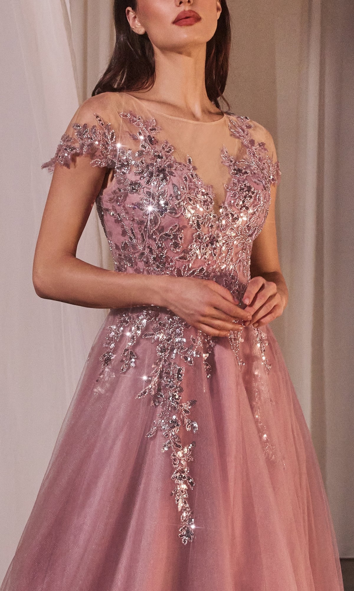 A woman wears the Dusty Rose Prom Ball Gown, Andrea and Leo A1350—featuring a long pink design, sheer cap sleeves, and sparkling silver floral embellishments—as she stands with her hands clasped.