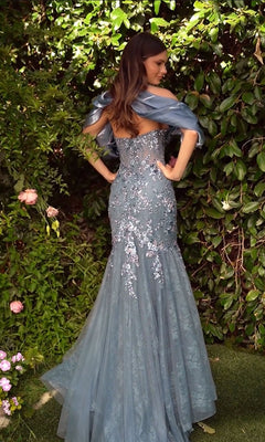 A woman stands in a garden, facing away, wearing the Andrea & Leo A1354 Long Mermaid Prom Dress—a fitted, strapless blue lace gown with floral embroidery.