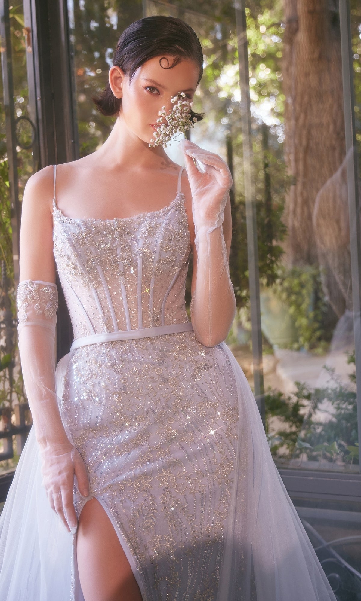A woman wearing the Andrea and Leo A1361 beaded light blue prom gown with sheer gloves holds a small bouquet near her face, standing by a window overlooking lush greenery.