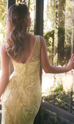 A woman in the Illusion Long Beaded Prom Gown by Andrea & Leo (A1362) stands by a window, facing outside with one hand on the glass, amid lush greenery.